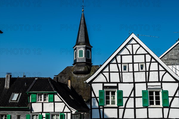 White half-timbered house with green shutters in front of a church tower under a blue sky, Graefrath, Solingen, Bergisches Land, North Rhine-Westphalia