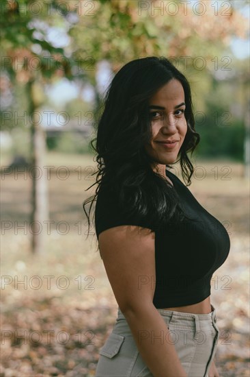 A smiling Cheerful hispanic young woman outdoors wearing a black shirt and beige pants with trees in the background, selective focus, blurred forest, with bokeh, daytime, AI generated