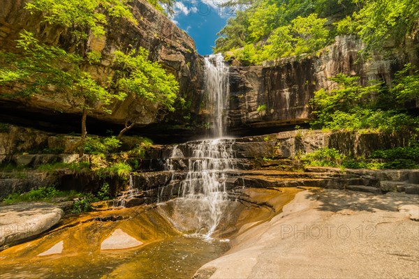 A tall waterfall plummets down a rocky cliff surrounded by vibrant green foliage, in South Korea