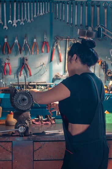 Woman mechanic repairing an alternator at a workbench in an auto moto workshop, wearing black overall and shirt, a complete tool panel in background with bokeh effect, traditional male jobs by Mixed-race latino woman
