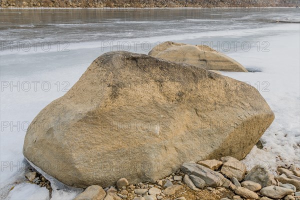 A large rock on the snowy bank of a partially frozen river, in South Korea