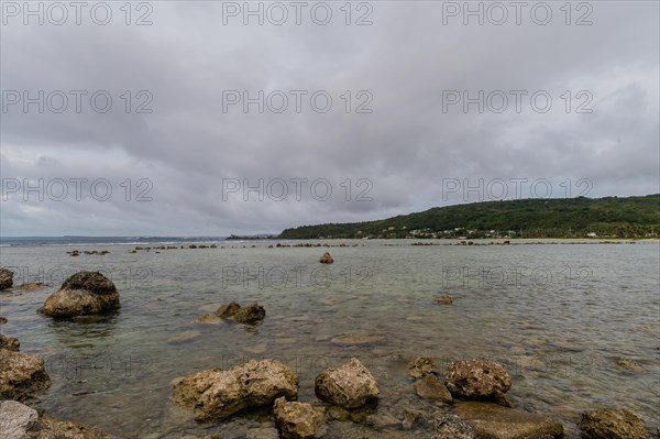 Seascape of rocky shoreline on a cloudy day with buildings on tree lined shore in the distance in Guam