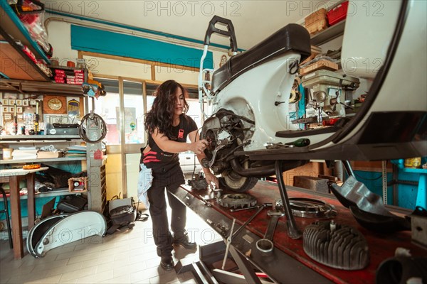 A woman mechanic works on a vintage scooter in a cluttered workshop, wearing black overall and shirt, latino female in traditional masculine jobs concept, feminine power in real life