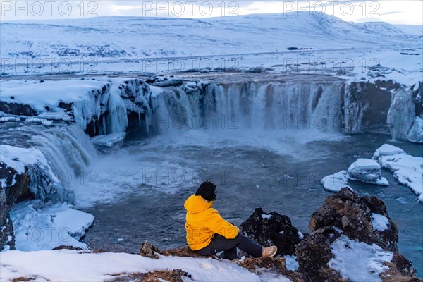 Adventurous woman in winter in Iceland sitting looking at frozen Godafoss waterfall