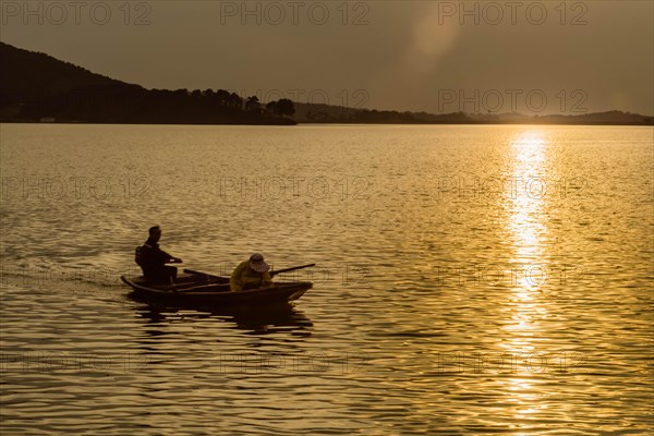 Two fishermen on a boat in a lake during sunset, framed by golden light and calm waters, in South Korea