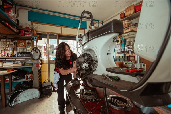 Determined woman mechanic inspecting the front of a white old scooter during an engine check, latino female in traditional masculine jobs concept, feminine power in real life