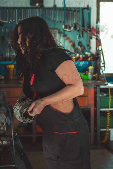 Young confident skilled hispanic Mechanic woman is closely examining a vehicle part in a garage setting, a complete tool panel in background with bokeh effect, traditional male jobs by Mixed-race latino female person