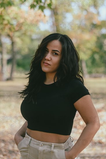 Cheerful hispanic young sensual Woman in a black shirt and beige pants outdoors with hands on hips and a serious expression, selective focus, blurred background with bokeh, daytime, AI generated