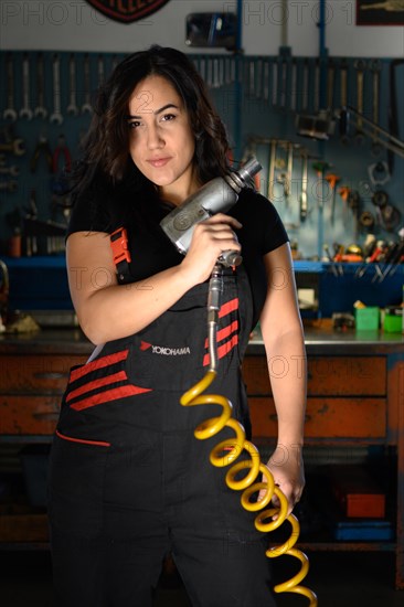 Empowered curvy hispanic sexy woman mechanic holding an air compressor hose in a dynamic pose in the garage, a complete tool panel in background with bokeh effect, traditional male jobs by Mixed-race latino woman