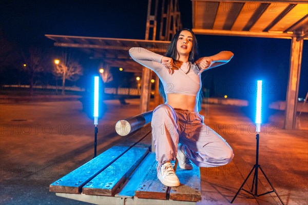 Confident trap artist posing crouching outdoors at night next to neon lights