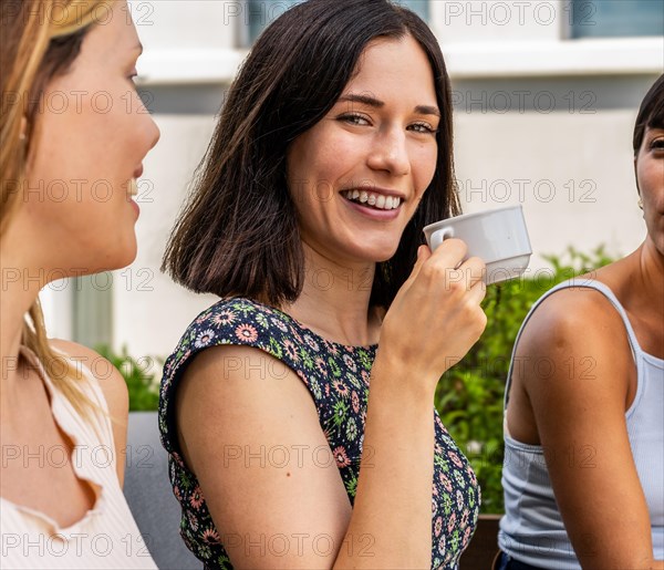 Three women are sitting together and one of them is holding a cup. The woman with the cup is smiling and looking at camera and the other two women are looking at her