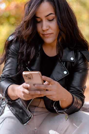 Young woman intensely focused on her phone, sitting in an autumn setting, blurred background with bokeh, daytime, AI generated