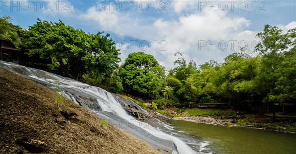 Side view of a waterfall cascading down a cliff into the basin below with green trees and blue, cloudy sky in the background in Guam