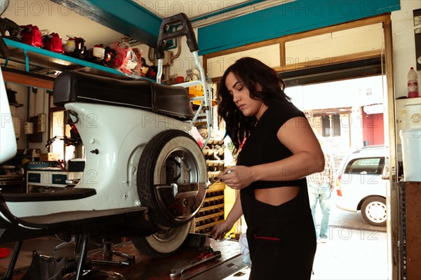 Sexy woman confident Mechanic in black attire fixing a tire on a white scooter in a workshop, latino female in traditional masculine jobs concept, feminine power in real life