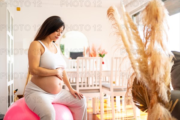 Pregnant woman touching her belly sitting on pink pilates ball in the living room after exercising