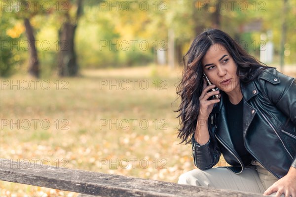 Cheerful hispanic young Woman engaged in a phone call while sitting on a park bench in autumn, blurred background with bokeh, daytime, AI generated