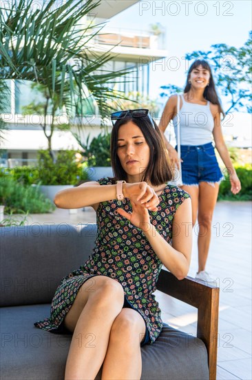 A woman is sitting on a couch and looking at her watch waiting for a friend who is behind her
