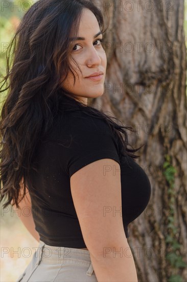 Cheerful hispanic young Woman in black shirt turning her head with a soft smile by a tree, blurred background with bokeh, daytime, AI generated