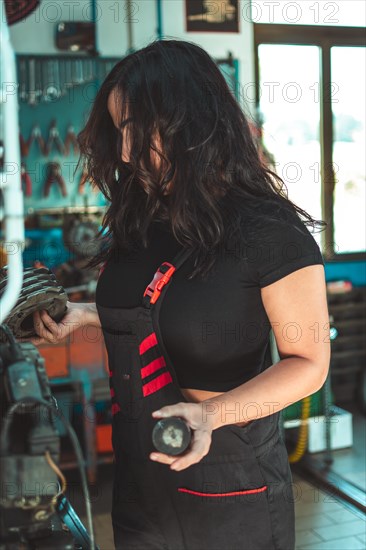 Young confident skilled hispanic long haired brunette woman in a mechanic's apron is examining a car part in a workshop, a complete tool panel in background with bokeh effect, traditional male jobs by Mixed-race latino female