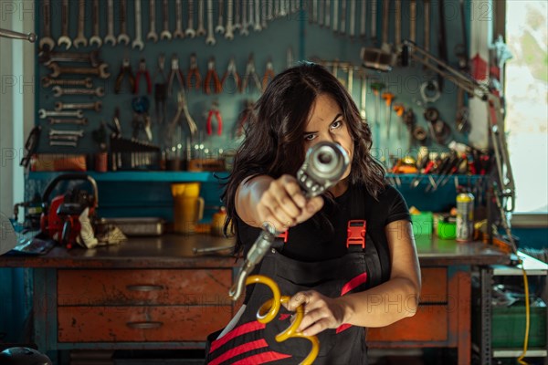 Intense sexy woman mechanic aiming a tool at the camera in a workshop, a complete tool panel in background with bokeh effect, traditional male jobs by Mixed-race latino woman