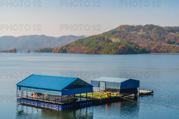 A floating structure with a blue roof on a calm lake with mountains in the distance, in South Korea