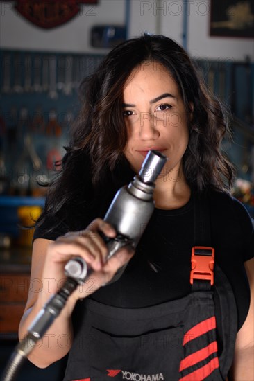 Playful confident woman mechanic holding an air hose and making eye contact in a workshop, a complete tool panel in background with bokeh effect, traditional male jobs by Mixed-race latino woman