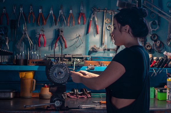 Mixed-race hispanic young Female mechanic, wear black overall, focused on an motorcycle part in a workshop with a tool cabinet, a complete tool panel in background with bokeh effect, traditional male jobs by Mixed-race latino woman