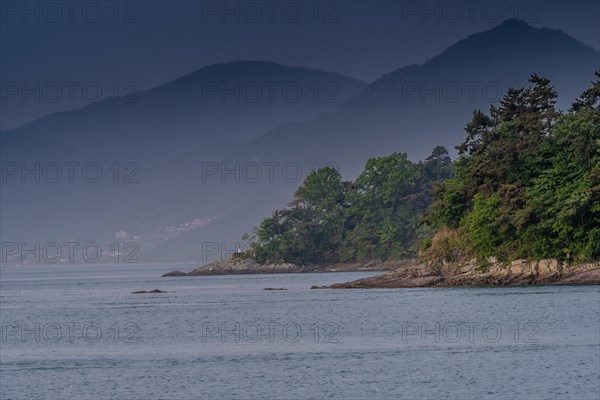 Landscape of shoreline with lush green trees and small mountains shrouded in fog and mist in the background in Namhae, South Korea, Asia