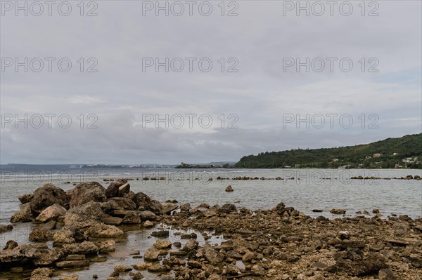 Seascape of rocky shoreline on a cloudy day with buildings on tree lined shore in the distance in Namhae, South Korea, Asia