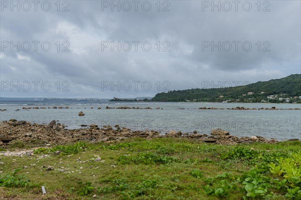 Seascape of rocky shoreline on a cloudy day with buildings on tree lined shore in the distance in Guam