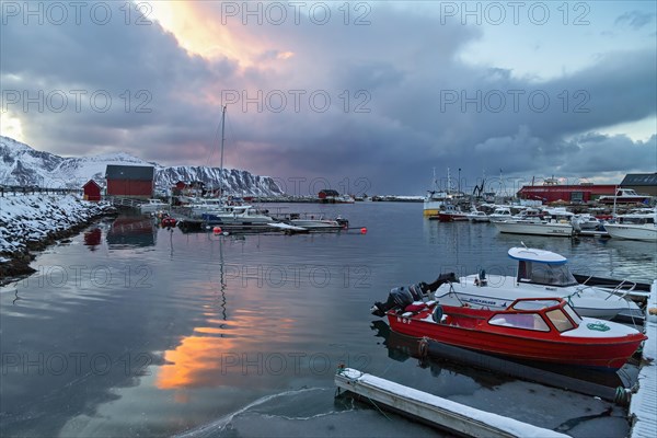 Twilight scene at a snow-covered harbor with boats and a vibrant sky reflection in the water, Lofoten