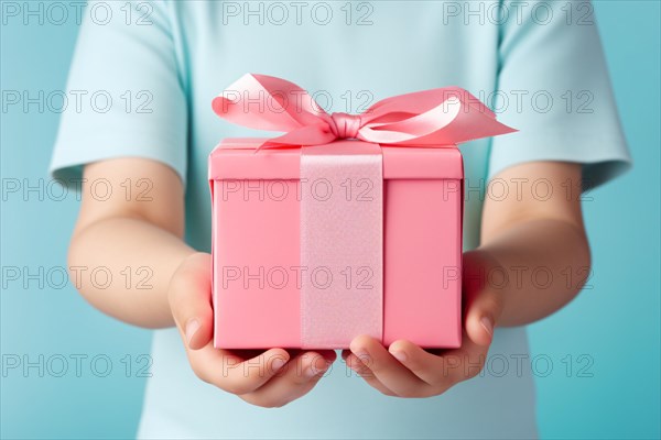 Child's hands holding pink Mother's day gift box with ribbon in front of pastel blue studio background. KI generiert, generiert AI generated