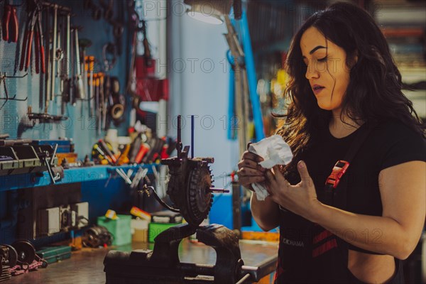 A female mechanic wearing overall in a workshop is carefully cleaning motorcycle parts, a complete tool panel in background with bokeh effect, traditional male jobs by Mixed-race latino woman