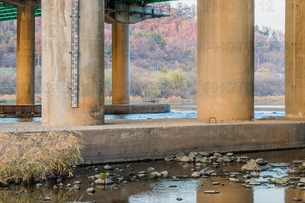 Pillars of a bridge over a river showing a water level gauge, with fall foliage around, in South Korea