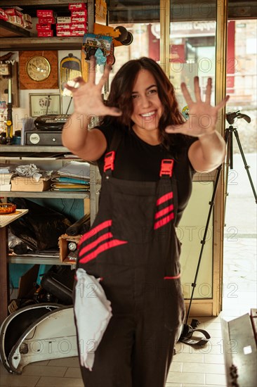 Cheerful hispanic young female mechanic wearing black overalls waving her hands in a workshop, latino woman in traditional masculine jobs concept, feminine power in real life