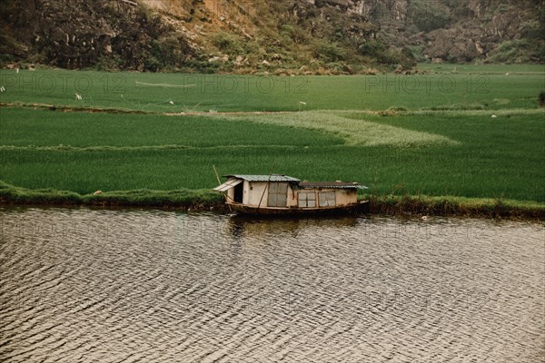 A small boat floats on a river with lush green fields and hills in the background, Ninh Binh, Vietnam, Asia