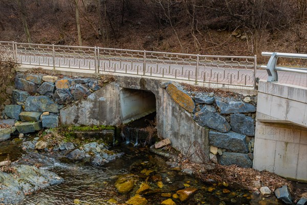 A small stream flows under a bridge with a stone wall and bare trees beside it, in South Korea