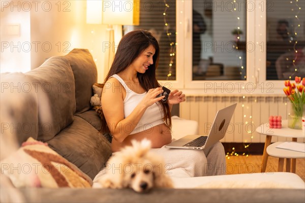 Pregnant woman using laptop sitting on the sofa to do a video call and showing a ultrasound from home