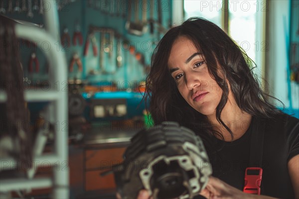 Woman mechanic holding a motorcycle engine part in a workshop, appearing thoughtful, a complete tool panel in background with bokeh effect, traditional male jobs by Mixed-race female professional