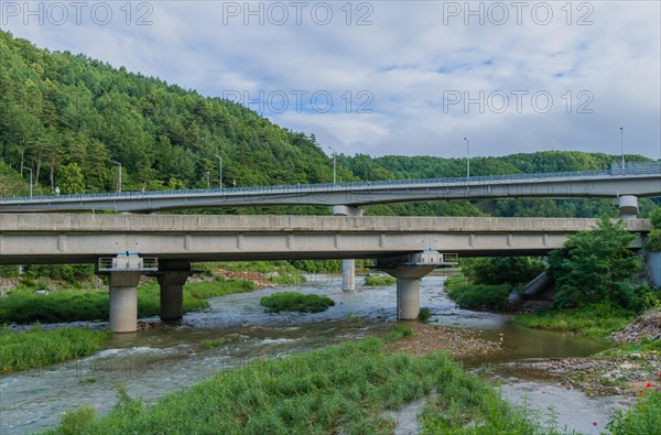 A concrete bridge over a river surrounded by green foliage under an overcast sky, in South Korea