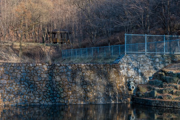 Pond enclosed by a stone wall with a barrier and bare trees reflecting in still water, in South Korea