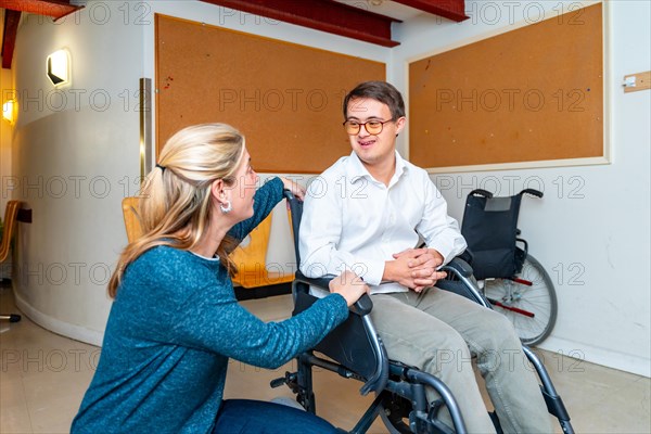 Kind female caregiver talking to a man with down syndrome on wheelchair in a day center