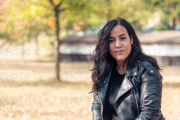 A cheerful hispanic young woman standing in a park with trees wearing a black rider leather jacket, outdoors in autumn, blurred background with bokeh, daytime, AI generated
