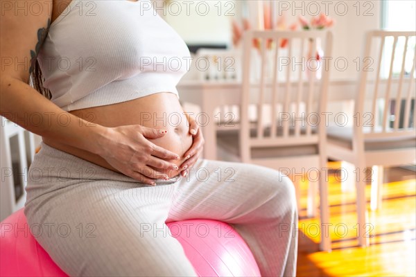 Unrecognizable pregnant woman touching her belly doing exercises with pilates ball at home