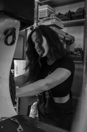 Determined woman mechanic working on the engine of a moped italian vintage scooter in a garage, real women performing traditional man jobs of the past, black and white photograph