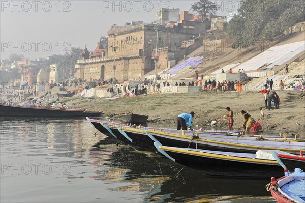 Lively scene by the river where boats lie on the banks and people walk on the ghats by the buildings, Varanasi, Uttar Pradesh, India, Asia
