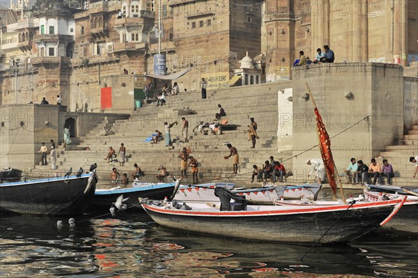 Boats on the bank of a river with people on stairs next to historical buildings, Varanasi, Uttar Pradesh, India, Asia