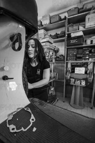 Serious woman mechanic disassembling a part on a moped italian vintage scooter motorcycle in the workshop, real women performing traditional man jobs of the past, black and white photograph