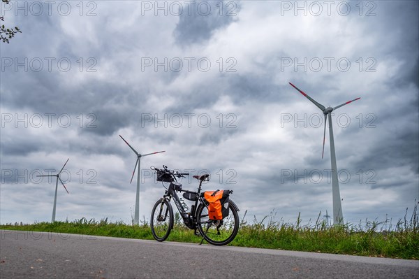 A parked bicycle at the roadside with wind turbines in the background under a cloudy sky, Leer, East Frisia, Lower Saxony