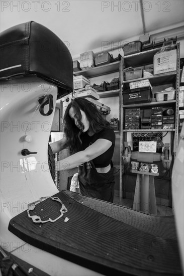 Woman mechanic focusing on repairing the inside of a moped italian vintage scooter in a workshop, real women performing traditional man jobs of the past, black and white photograph